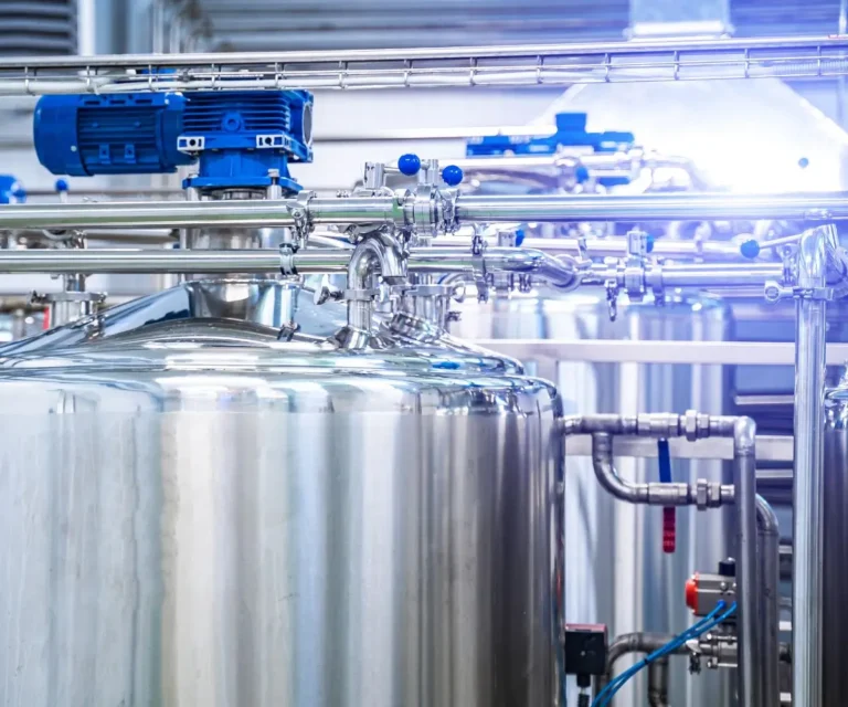 Jak technologia zmienia branżę mleczarską 3 Na zdjęciu znajduje się fragment systemu produkcyjnego w mleczarni. Centralną część obrazu zajmuje duży, błyszczący zbiornik ze stali nierdzewnej, z zamontowanymi na górze rurami i zaworami. Na szczycie widoczny jest niebieski silnik elektryczny, który najpewniej napędza mieszadło wewnątrz zbiornika. Wokół zbiornika znajdują się liczne metalowe rurki i połączenia, tworzące złożony układ, zapewniający transport cieczy lub gazów w różnych kierunkach. Zdjęcie jest dobrze oświetlone, a odbicia na metalowych powierzchniach dodają przestrzeni technicznego, sterylnego charakteru.